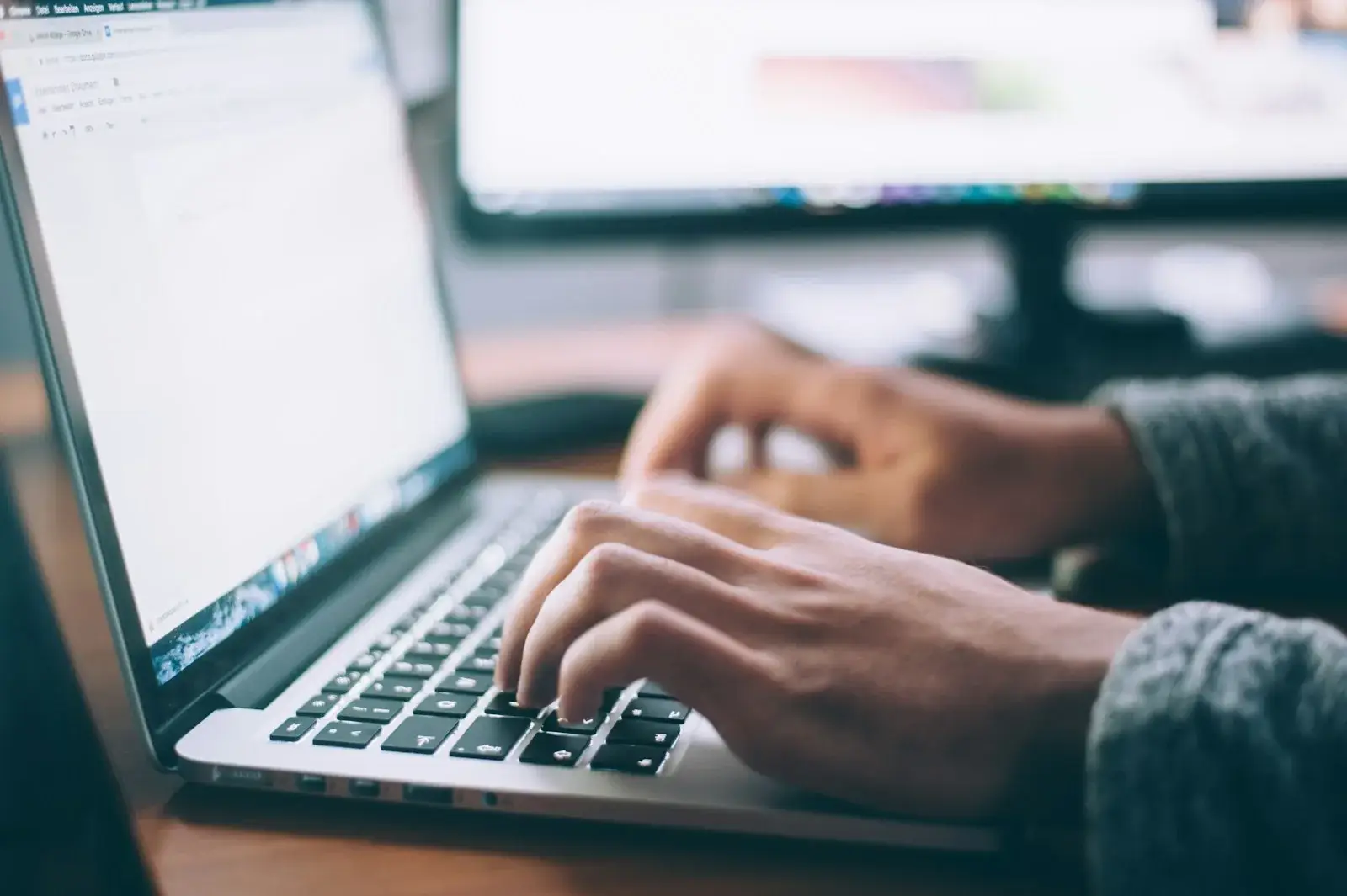 Hands typing on a laptop keyboard beside a desktop monitor
